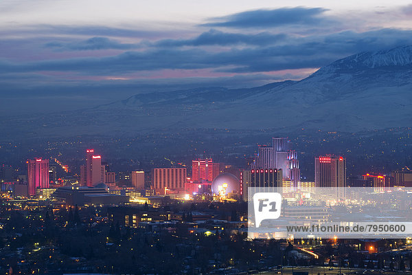 The Reno skyline is illuminated at dusk with clouds in the sky and snow covered peaks in the background in Reno  NV.