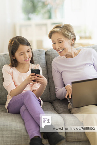 Granddaughter (8-9) and grandmother sitting on sofa and using laptop and cell phone