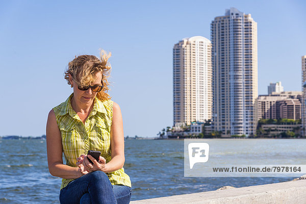 USA  Florida  Miami  Reife Frau  die auf der umgebenden Wand des Hafens sitzt und Smartphone benutzt.