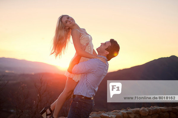 Junger Mann hebt bei Sonnenuntergang seine Freundin hoch.