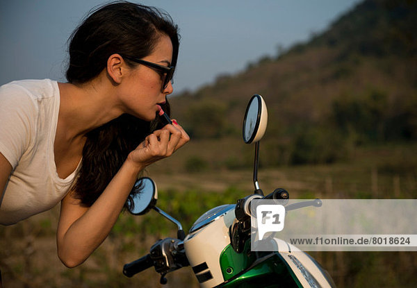 Woman applying lipstick in wing mirror of moped
