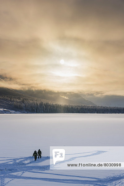 Sillhouetted ice fisherman on Summit Lake  clouds clearing from the Kenai Mountains behind the lake  Chugach National Forest  Backlit sun  Winter  Southcentral Alaska  USA.