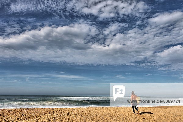 Seignosse le Penon (40). Surfer stripped to the waist on the beach  about to enter the water with his surfboard under his arm  on the Atlantic Coast. Landes department or moor  Surfing  Waves  rollers  Ocean  sand  blue sky  nice weather
