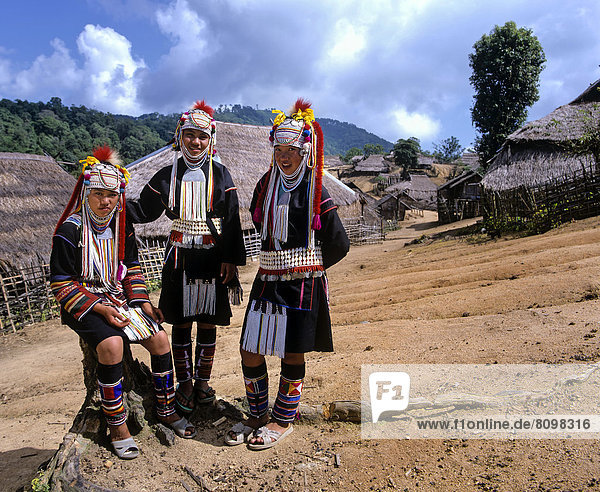 Three Akha girls in a mountain village  in traditional costume and headdress  bamboo huts with thatched roofs at back