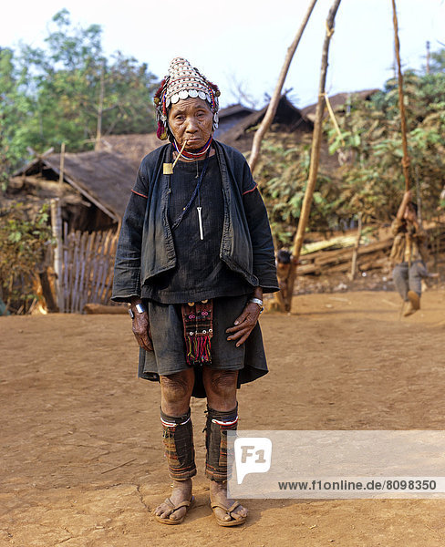 Elderly woman with a whistle standing in front of bamboo huts in an Akha village