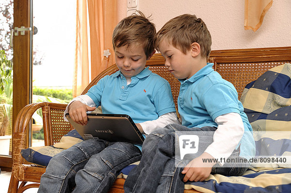 Two boys  twins  sitting side by side on a bench in a living room playing on a tablet computer