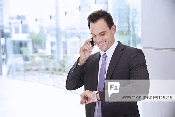 Smiling businessman talking on cell phone and looking at wristwatch