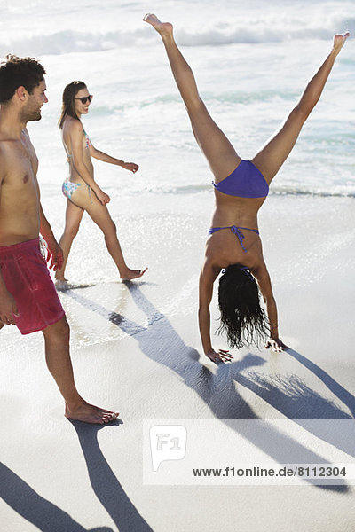 Freunde beobachten Frau im Bikini beim Handstand am Strand