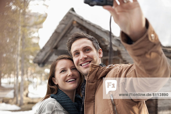 Smiling couple taking self-portrait with digital camera outside snowy cabin