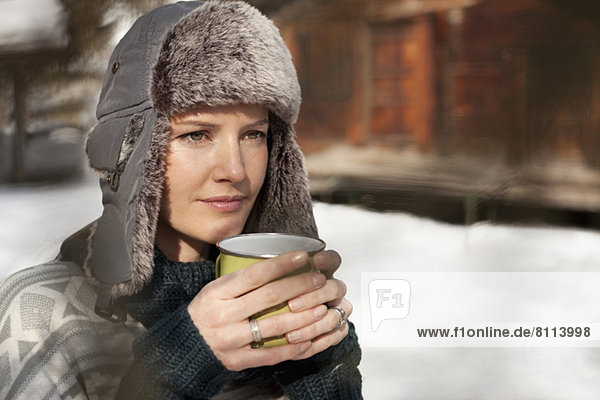 Pensive woman in fur hat drinking coffee outside cabin