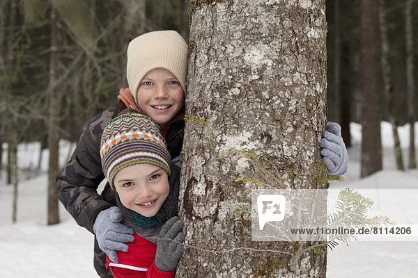 Portrait of happy boys behind tree trunk in snowy woods