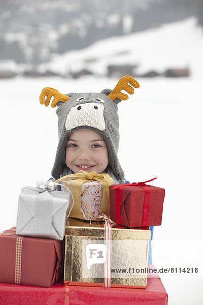 Portrait of smiling boy wearing reindeer hat and carrying stack of Christmas gifts in snow