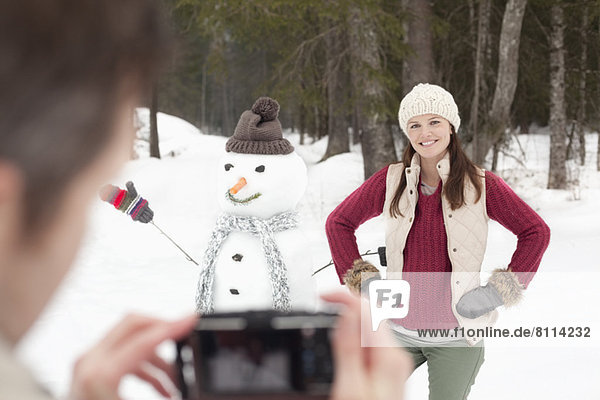 Man photographing smiling woman with hands on hips next to snowman in woods