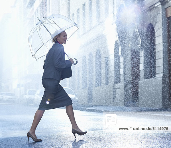 Glückliche Geschäftsfrau mit Regenschirm,  die über die regnerische Straße läuft.