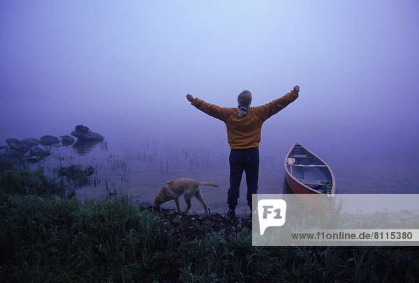 North America USA Maine Water Lake River Shore Canoe Arms Grass Stones Fog Mist Reflection One Dog One Person One Lady Standing Stretching Relaxing Refreshing Peaceful Fitness Exercise Outdoors Morning Foggy Rear View Horizontal Color Image Photography