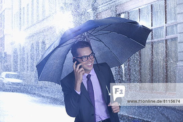 Happy businessman talking on cell phone under umbrella in rainy street