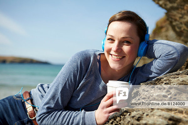 Porträt einer jungen Frau an der Küste mit Kaffee und Kopfhörer