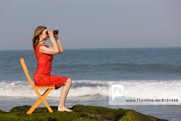 Reife Frau sitzt auf einem Stuhl am Strand mit Fernglas