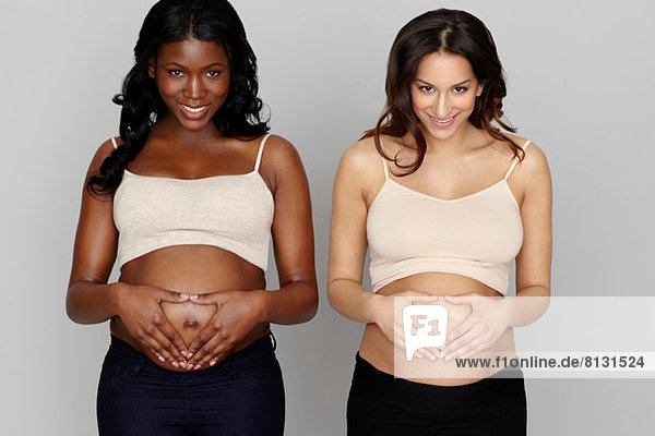 Zwei schwangere Frauen machen Herzformen mit Händen auf dem Bauch.