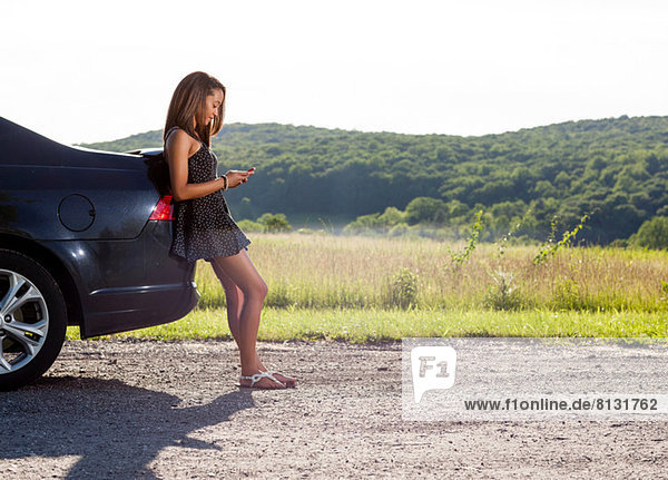 Junge Frau  die sich mit dem Handy gegen das Auto lehnt.