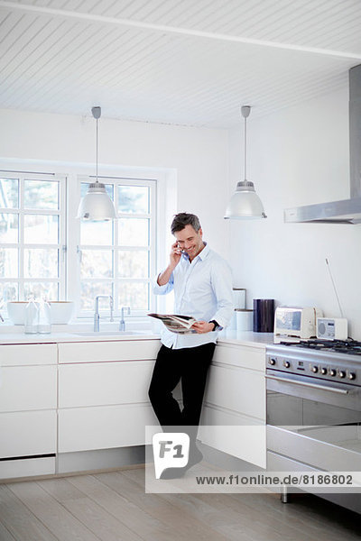 Mann steht in der Küche,  telefoniert und liest Zeitung.