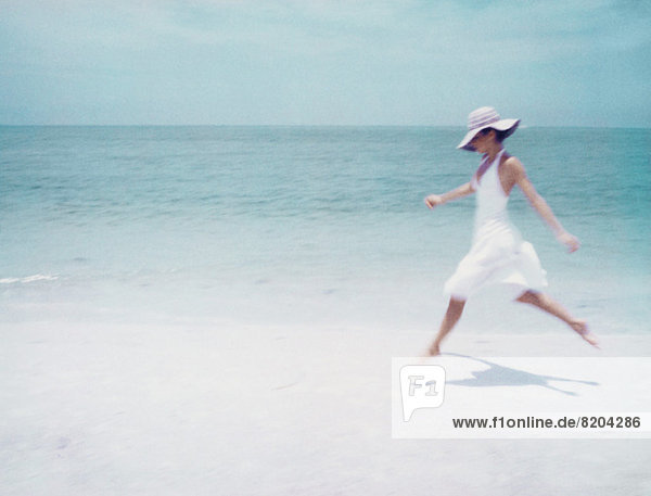 Junge Frau in Kleid und Sonnenhut  Laufen am Strand  Seitenansicht
