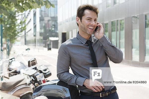 Businessman talking on cell phone in city  leaning against motor scooters