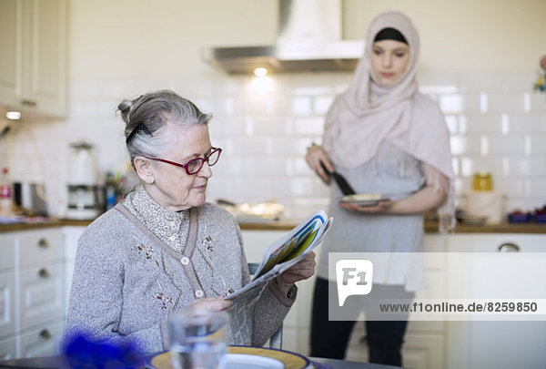 Seniorin liest Zeitung mit weiblicher Betreuerin  die in der Küche Essen zubereitet.