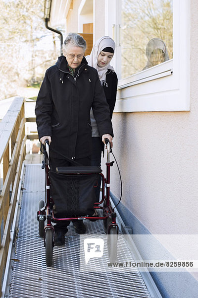 Heimpflegerin hilft Seniorin mit Gehgestell durch den Gang