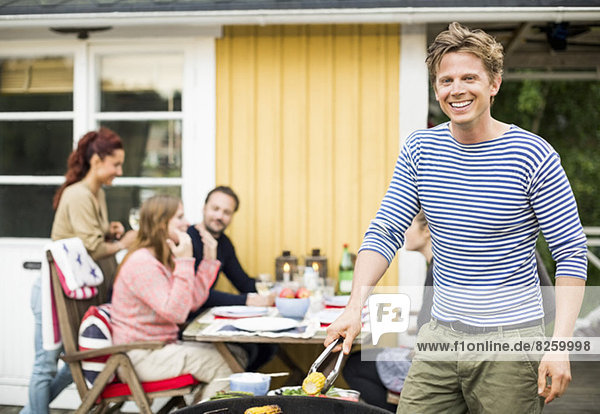 Porträt des glücklichen Mannes beim Grillen mit Freunden am Esstisch im Hintergrund