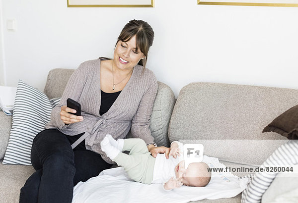 Mother using mobile phone while sitting with baby girl on sofa at home