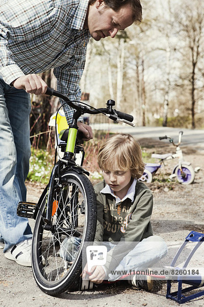 Junge füllt Fahrradreifen mit Fußpumpe  während Vater ihn auf der Straße beobachtet.