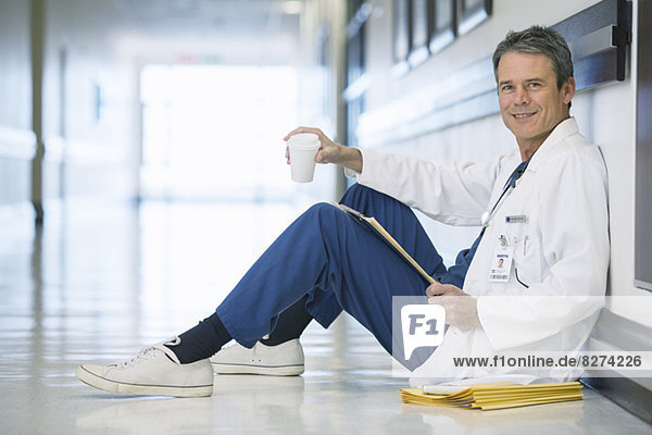 Porträt eines lächelnden Arztes beim Kaffeetrinken auf dem Boden im Krankenhausflur