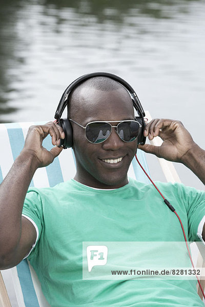 Man relaxing on deck chair by lake wearing headphones