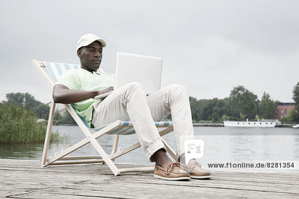 Man relaxing on deckchair with laptop by lake