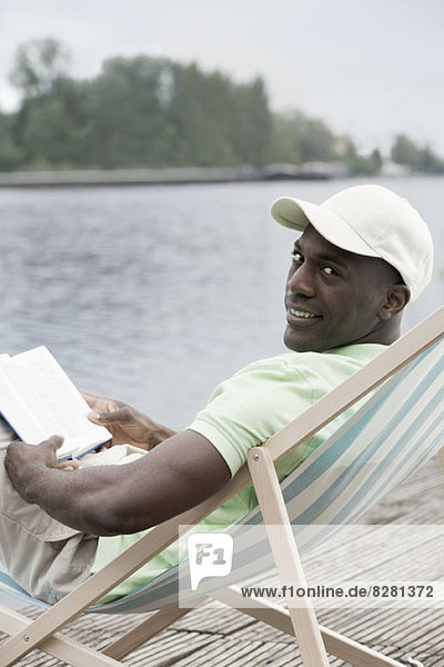 Man relaxing with book on deck chair by lake