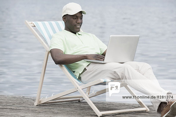 Man relaxing on deck chair using laptop by lake