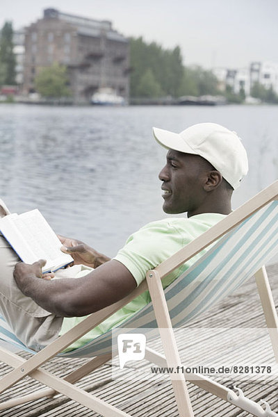 Man relaxing with book on deck chair by lake