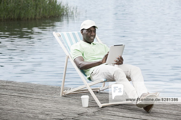 Man relaxing on deck chair using digital tablet by lake