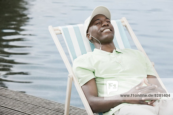 Happy man relaxing on deck chair by lake wearing headphones