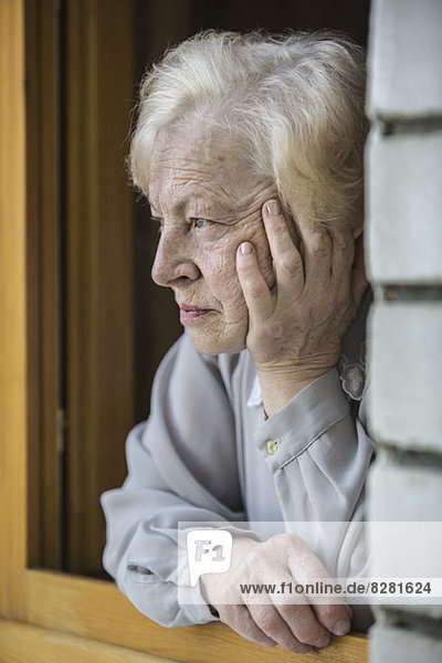 Eine ältere Frau  die sich auf ein Fensterbrett stützt und kontemplativ aussieht.