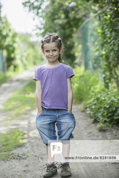 Ein junges lächelndes Mädchen mit Händen in den Taschen  das auf einem Feldweg steht.