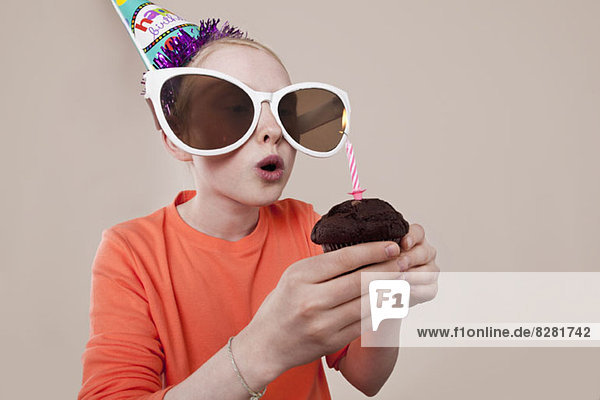 Ein Mädchen mit neuartiger Sonnenbrille und Partyhut beim Ausblasen der Geburtstagskerze