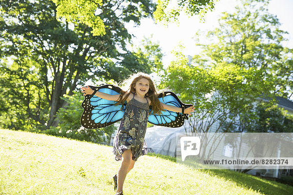 Ein Kind rennt vor einem Bauernhaus über den Rasen  mit großen reißenden blauen Schmetterlingsflügeln und mit ausgestreckten Armen.