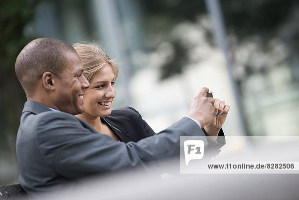 Eine junge blonde Frau und ein Mann auf einer Straße in New York City. Trägt Geschäftskleidung. Stehen Seite an Seite und fotografieren sich selbst mit einem Smartphone.