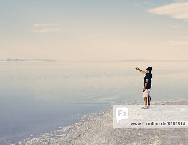 A Man Standing At Edge Of The Flooded Bonneville Salt Flats At Dusk  Taking A Photograph With A Smart Phone