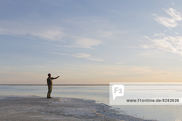 A Man Standing At Edge Of The Flooded Bonneville Salt Flats At Dusk  Taking A Photograph With A Tablet Device.