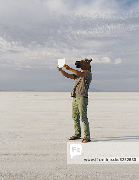 Ein Mann  der eine Pferdemaske trägt und mit einem Tablettengerät fotografiert  auf den Bonneville Salt Flats.