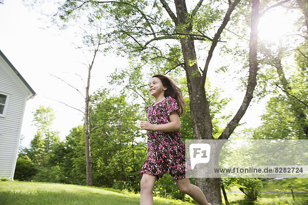 Ein junges Mädchen in einem gemusterten Sommerkleid  das unter dem Schatten der Bäume in einem Bauerngarten über das Gras rennt.