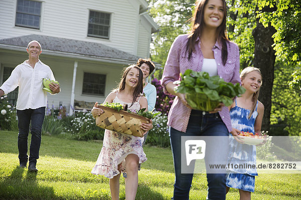 Familienfeier. Eltern und Kinder  die mit Blumen  frisch gepflücktem Gemüse und Obst über den Rasen laufen. Vorbereitungen für eine Party.